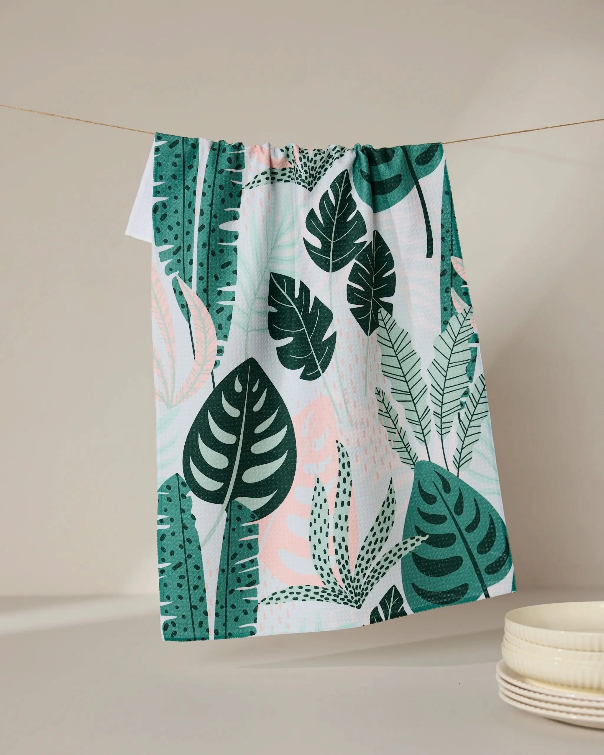The Diedre tea towel by GEOMETRY, featuring a quick-drying green and pink tropical leaf pattern, hangs on a string against a neutral background with white dishes stacked in the lower right corner.