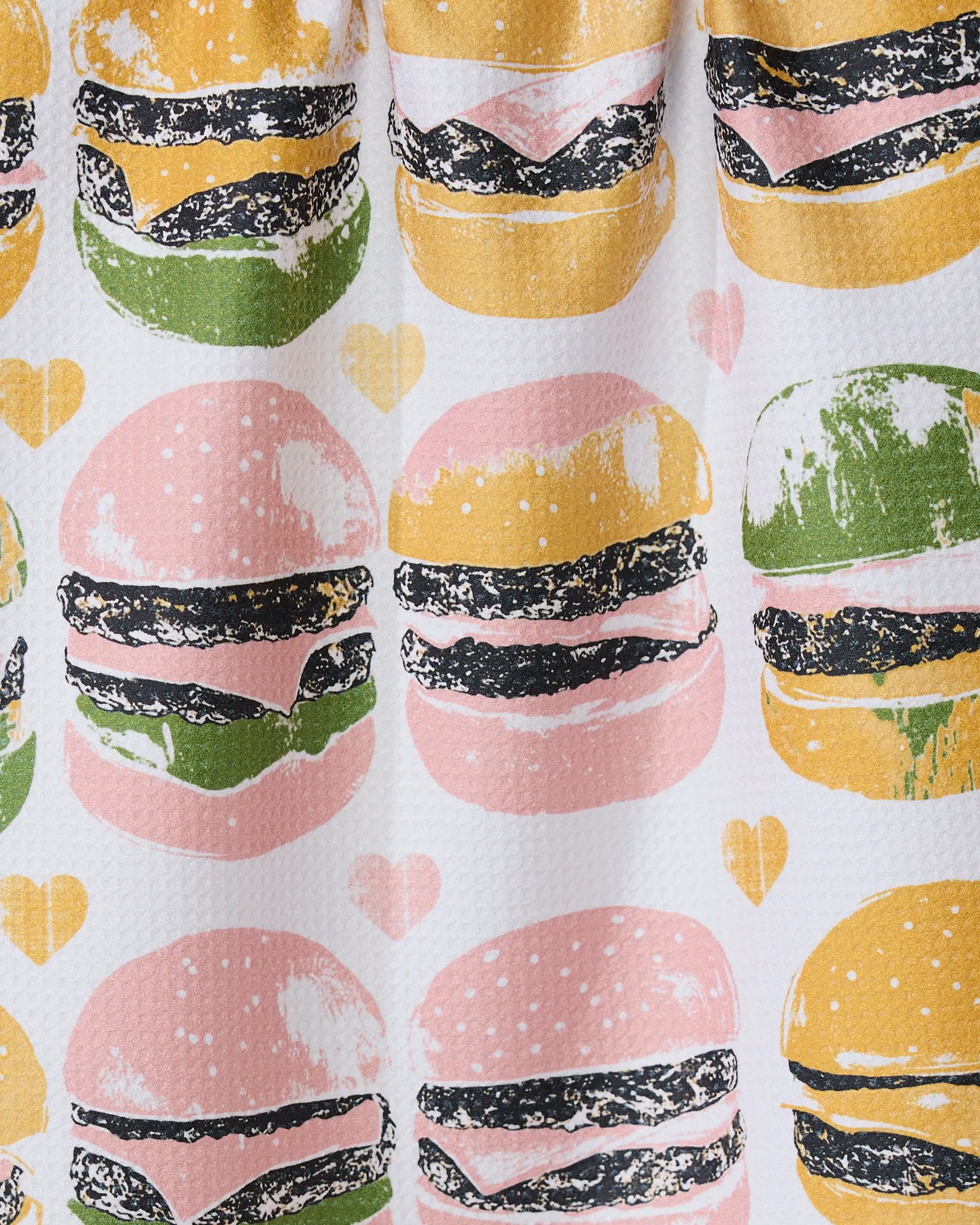 Geometry’s On The Grill tea towel features repeated burger illustrations with pink, yellow, and green buns plus small yellow and pink hearts on a white background.