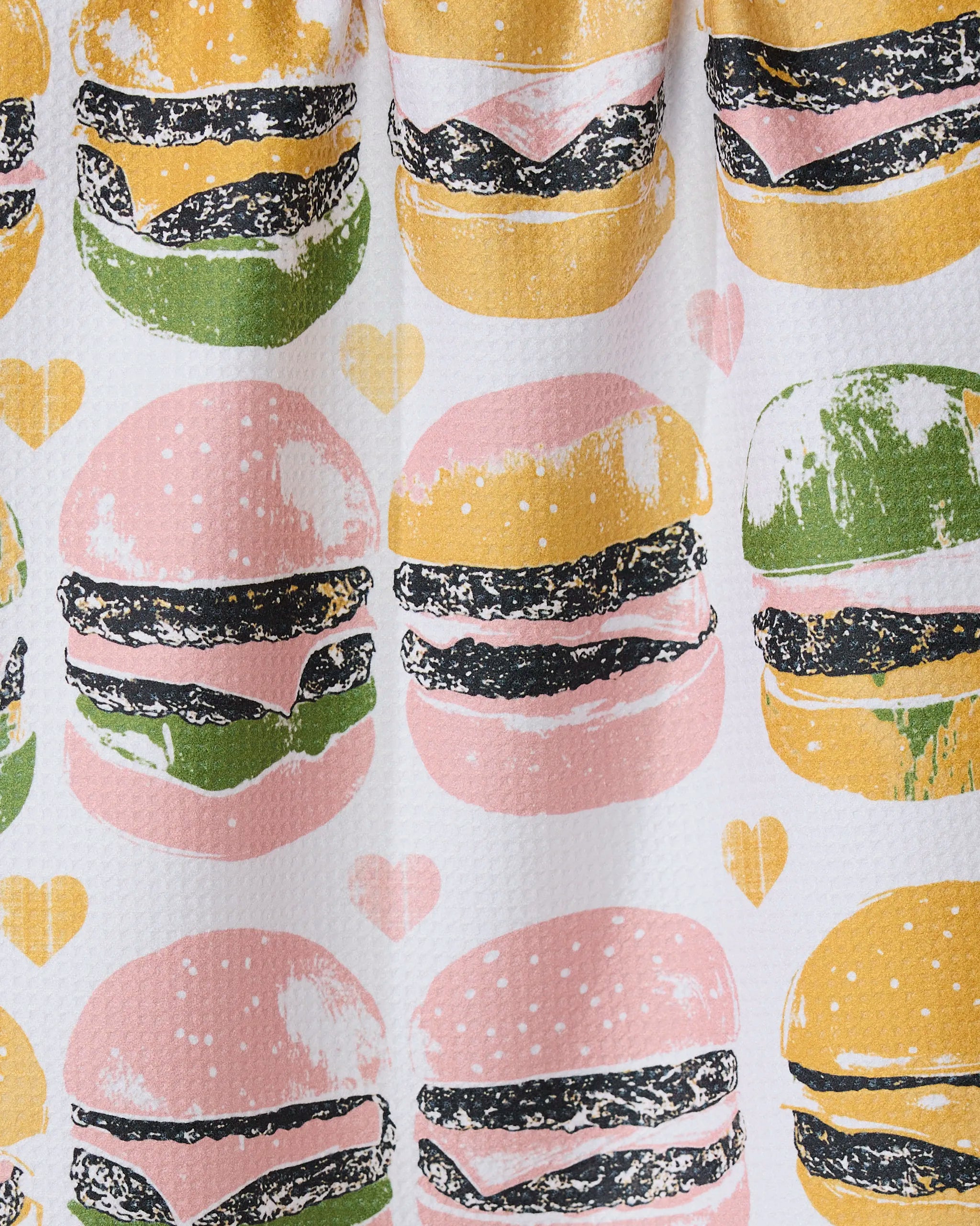 Geometry’s On The Grill tea towel features repeated burger illustrations with pink, yellow, and green buns plus small yellow and pink hearts on a white background.
