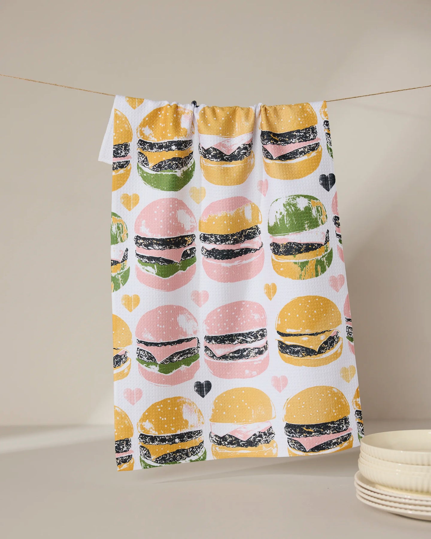 The Geometry On The Grill tea towel, featuring a vibrant hamburger and heart pattern, hangs above stacked white plates against a neutral backdrop.