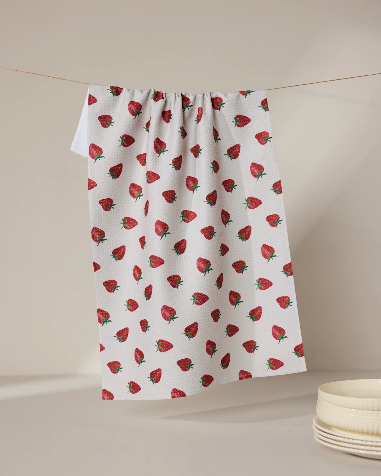 The Geometry Berry Patch tea towel, featuring a red strawberry pattern, hangs against a neutral backdrop beside stacked white bowls. Ultra-absorbent and stylish, it blends function with charm for any kitchen setting.