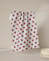 The Geometry Berry Patch tea towel, featuring a red strawberry pattern, hangs against a neutral backdrop beside stacked white bowls. Ultra-absorbent and stylish, it blends function with charm for any kitchen setting.