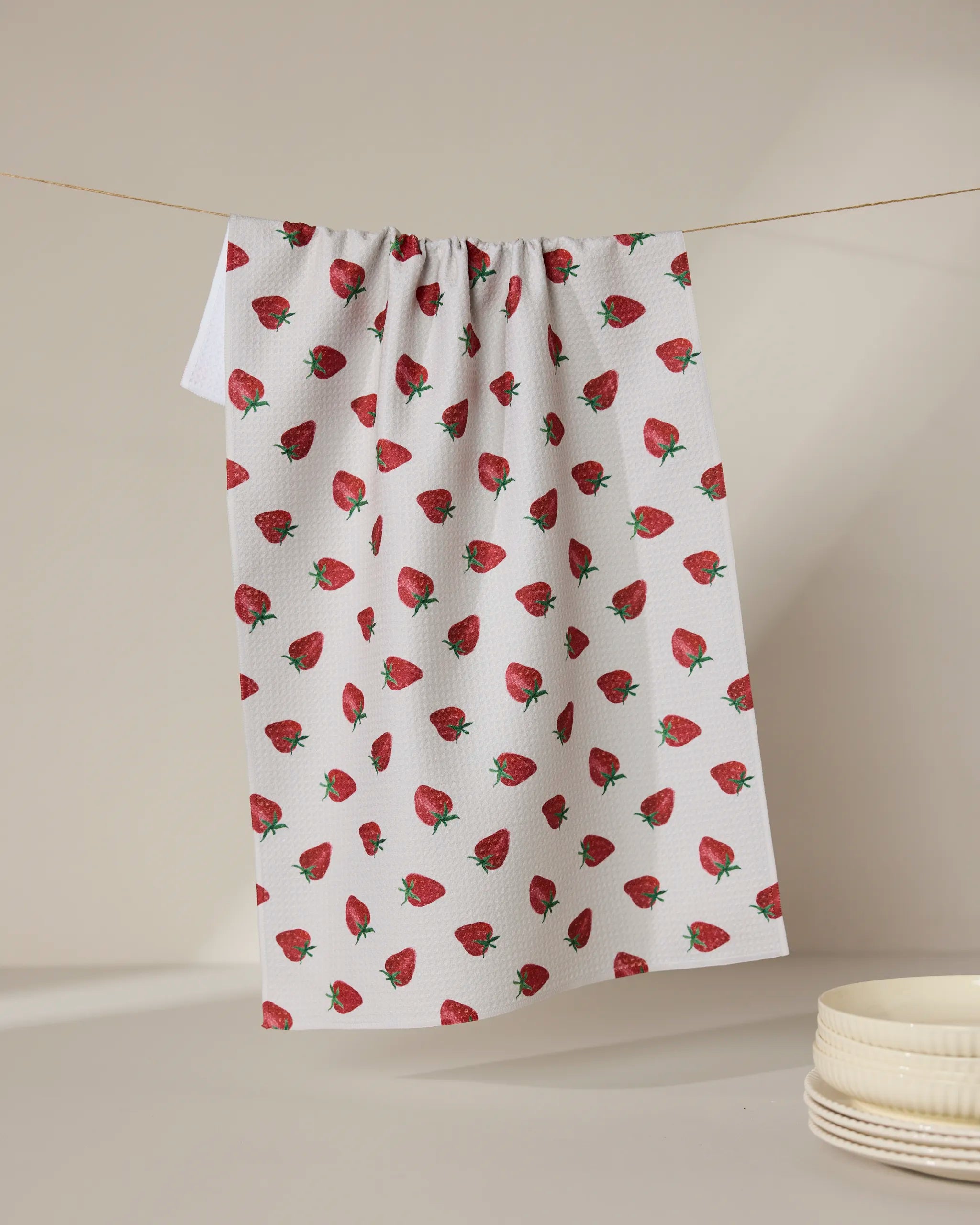 The Geometry Berry Patch tea towel, featuring a red strawberry pattern, hangs against a neutral backdrop beside stacked white bowls. Ultra-absorbent and stylish, it blends function with charm for any kitchen setting.