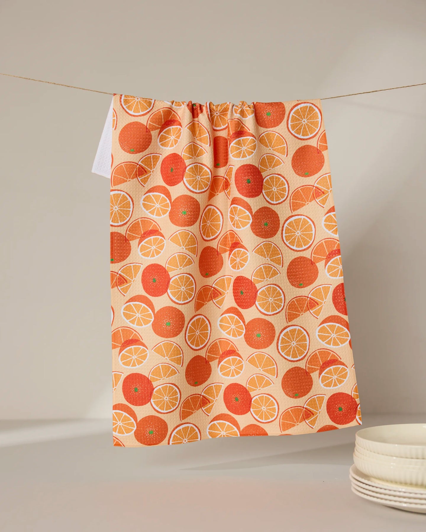 The Geometry Only Oranges recycled kitchen towel, featuring a vibrant orange slice and whole orange pattern, hangs on a clothesline with a stack of white dishes partially visible in the bottom right corner.