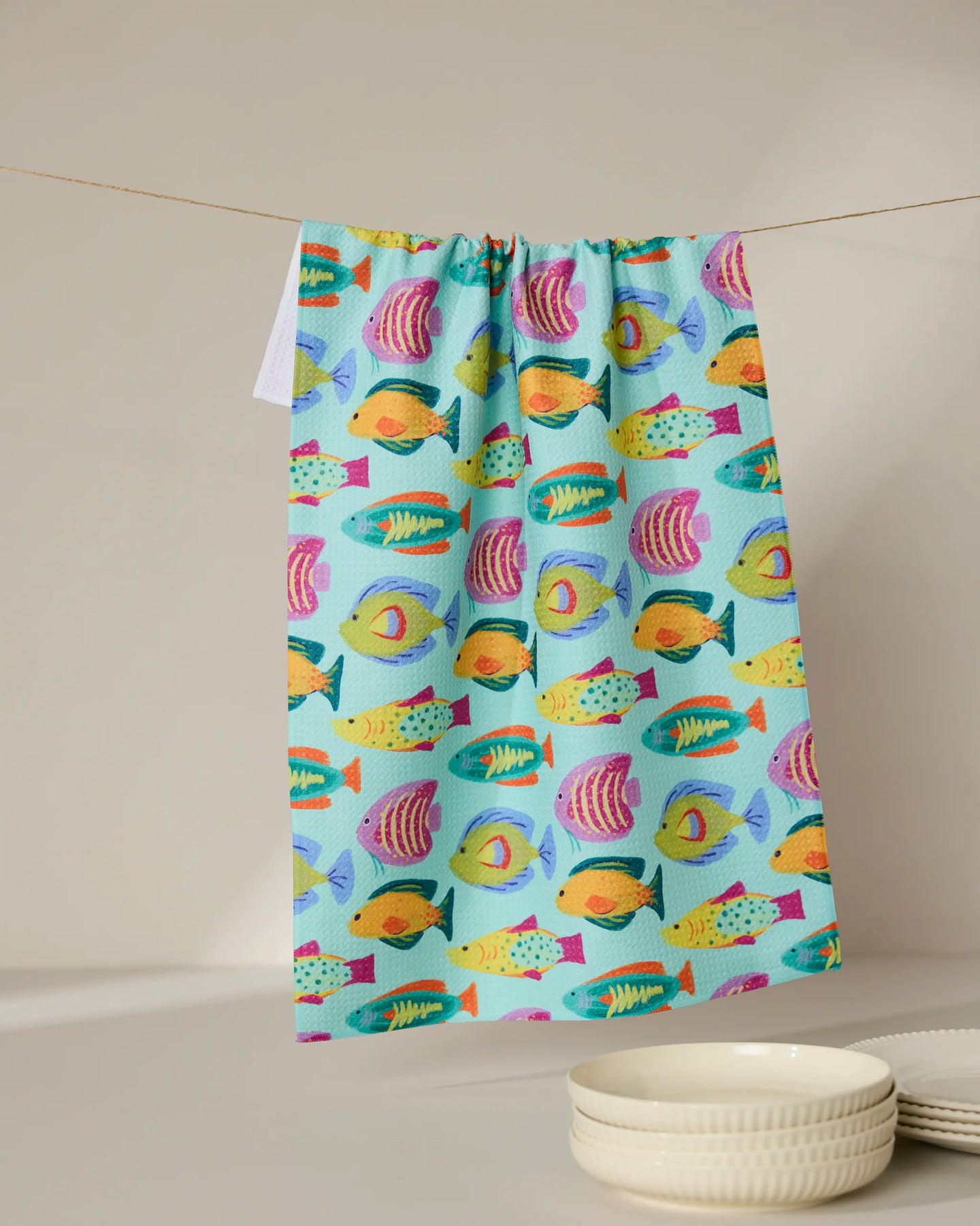 Geometrys Schooling kitchen towel, featuring a playful fish pattern and made from recycled materials, hangs above stacked white bowls against a neutral backdrop.