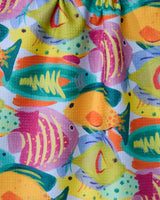 Geometrys Shoaling towel features a vibrant pattern of illustrated tropical fish in yellow, orange, green, pink, and purple overlapping on a white background. This colorful towel is quick-drying and perfect for summer days.