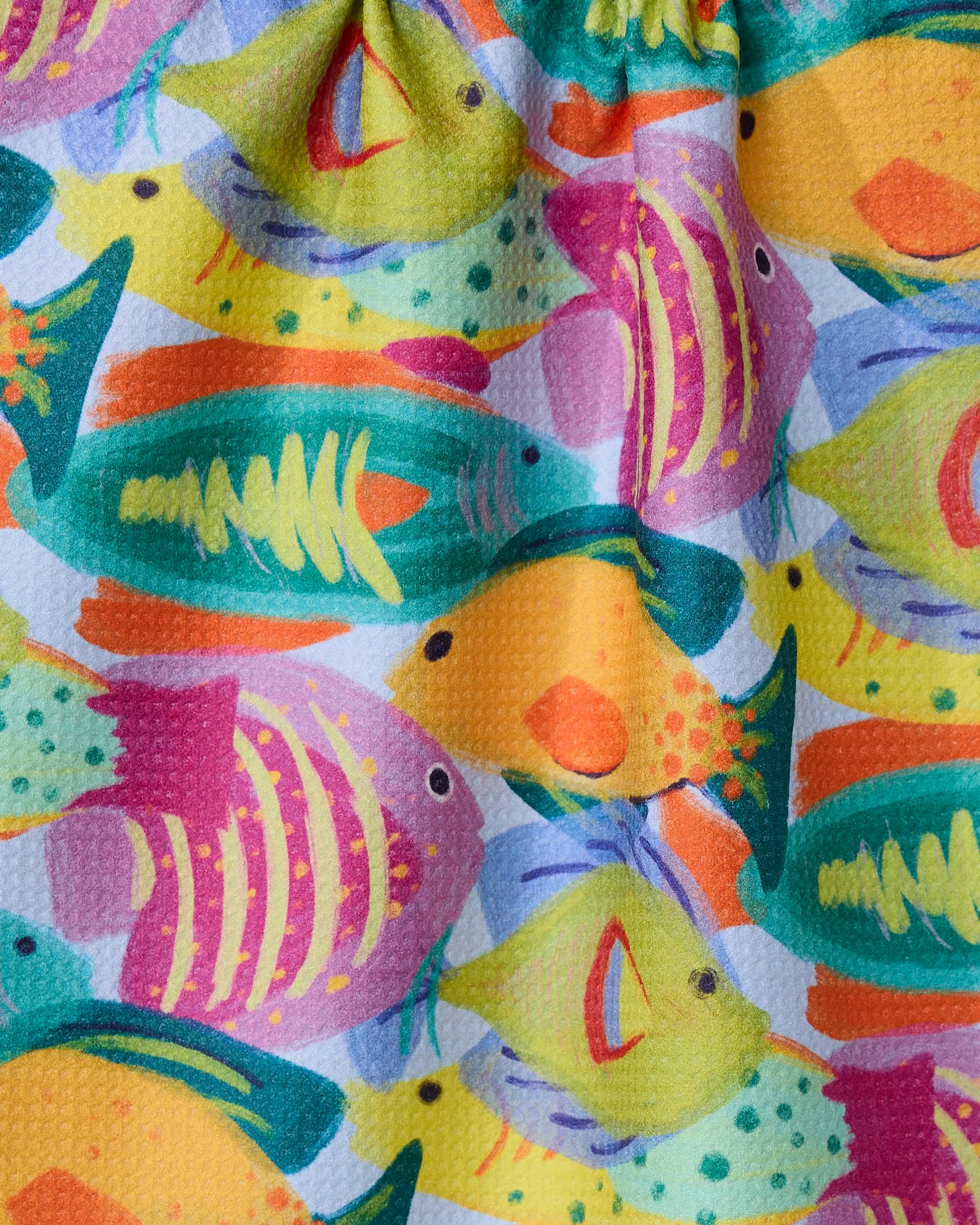 Geometrys Shoaling towel features a vibrant pattern of illustrated tropical fish in yellow, orange, green, pink, and purple overlapping on a white background. This colorful towel is quick-drying and perfect for summer days.