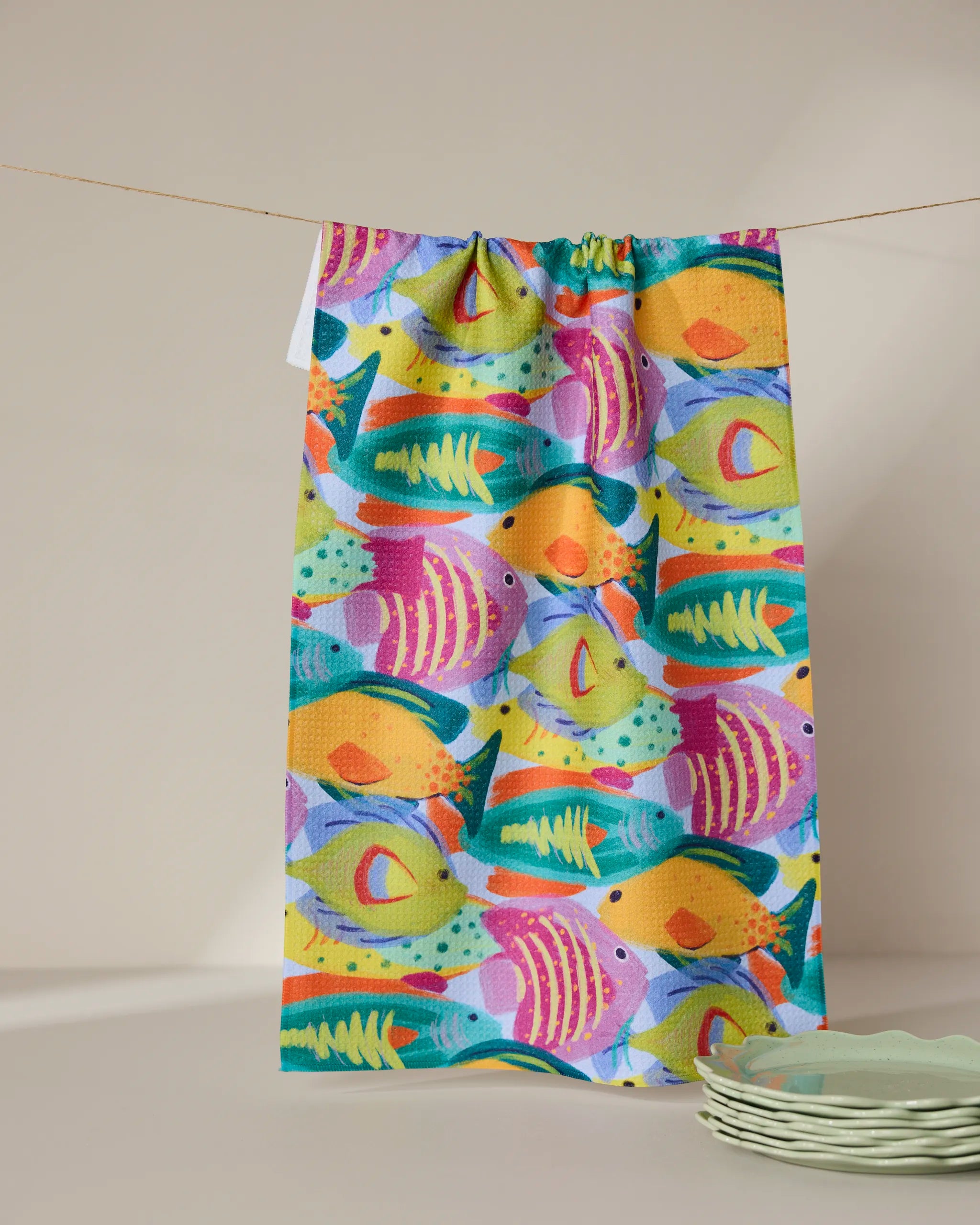 The Geometry Shoaling eco-friendly kitchen towel, featuring a vibrant fish pattern, hangs from a string against a neutral backdrop with green plates stacked nearby.