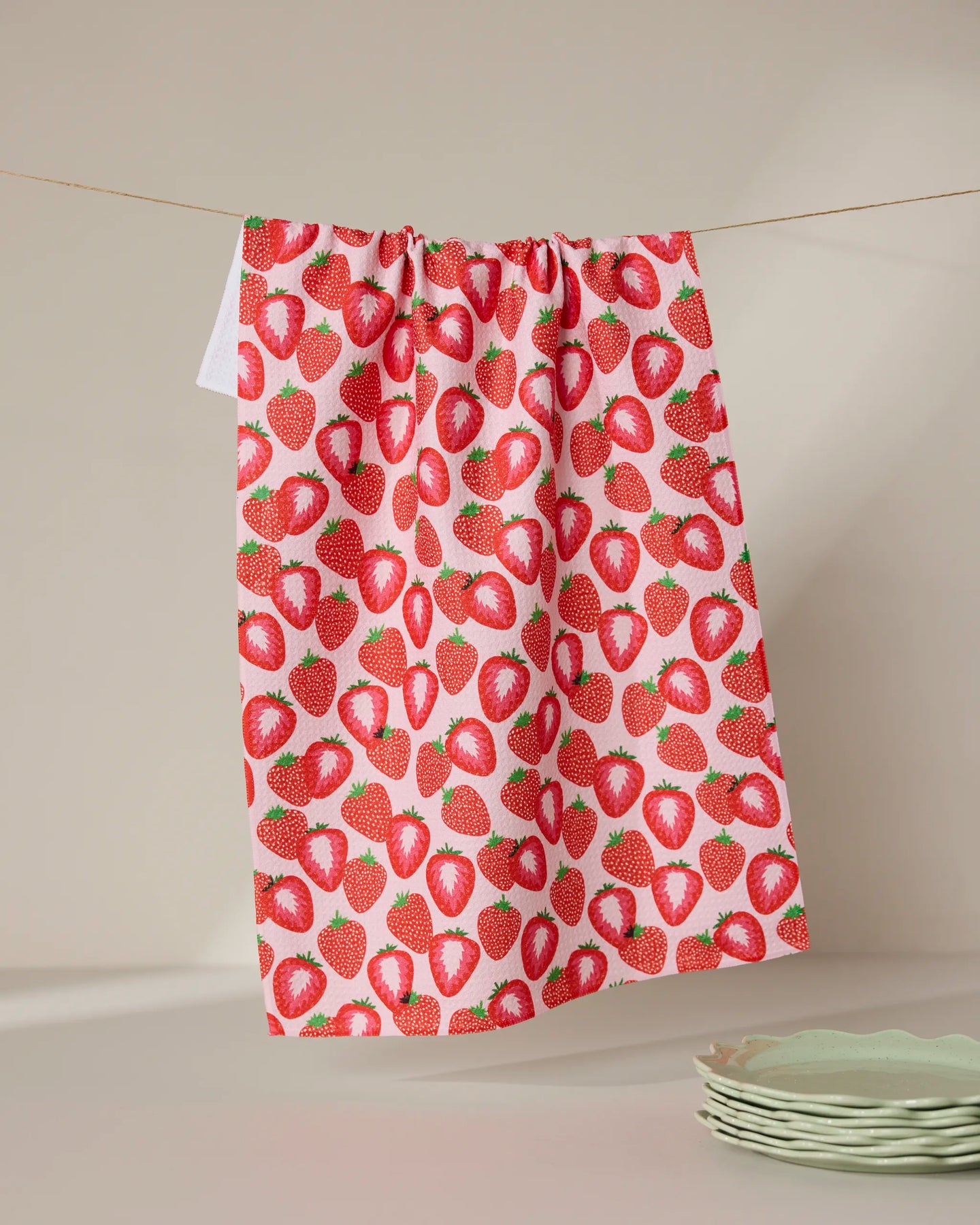 The Geometry So Many Strawberries tea towel, made from quick-drying recycled materials, hangs on a clothesline against a neutral background, with light green scalloped plates stacked in the lower right corner.
