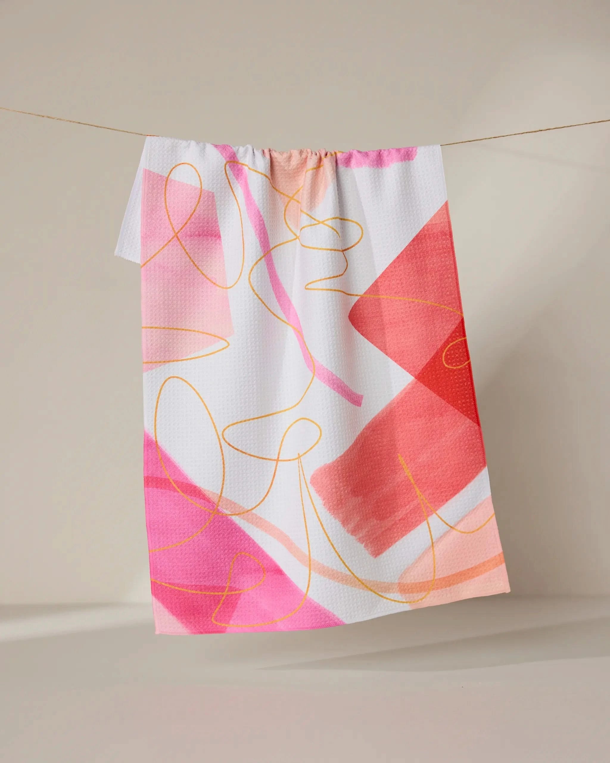The GEOMETRY Memories recycled kitchen towel, featuring bold abstract brushstrokes and swirling lines in pink, red, orange, and yellow, hangs on a string against a neutral background.