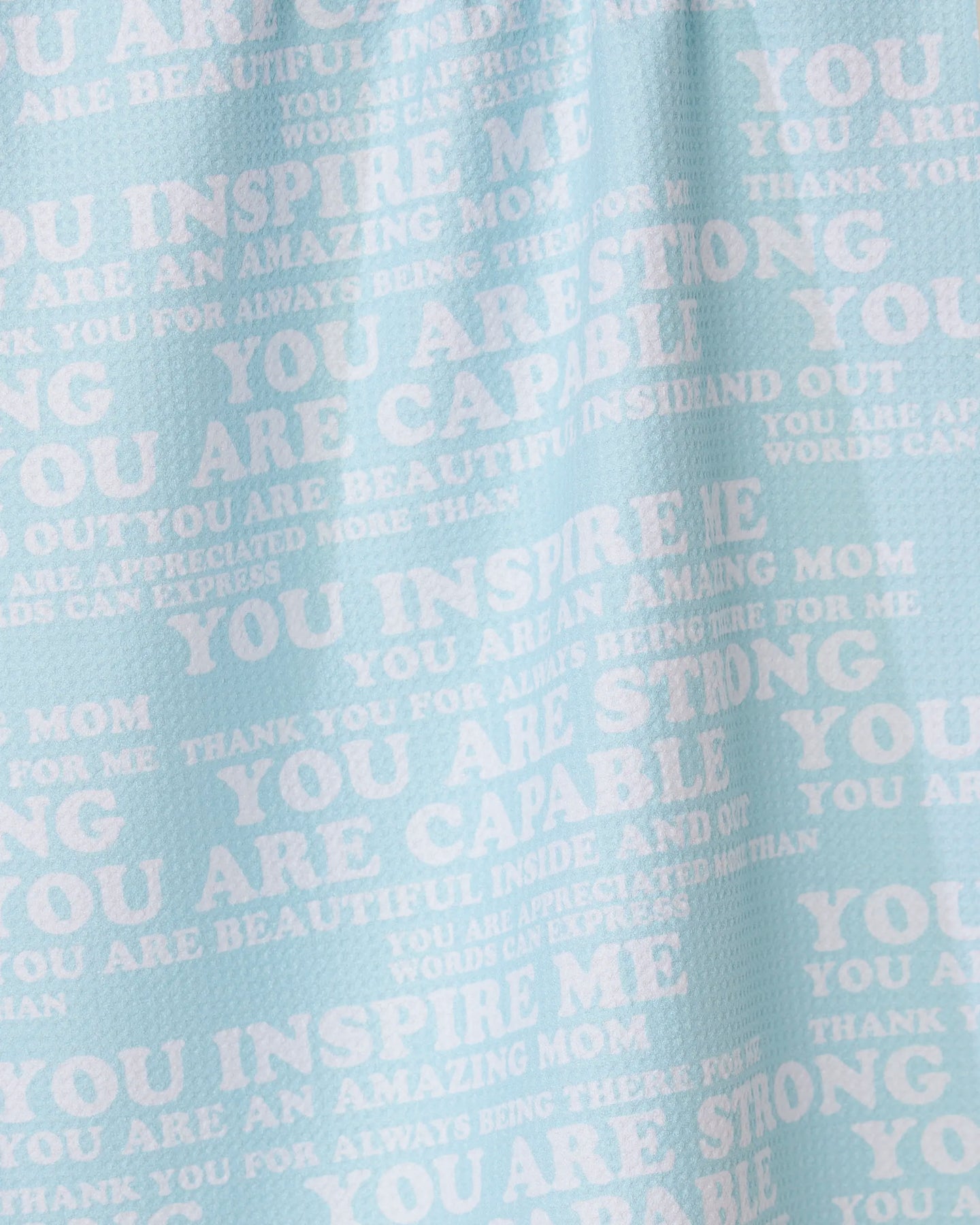 The GEOMETRY Mom Mantra tea towel is light blue, made from recycled materials, and features white overlapping motivational phrases like YOU INSPIRE ME and YOU ARE AMAZING MOM in different fonts and sizes.