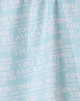The GEOMETRY Mom Mantra tea towel is light blue, made from recycled materials, and features white overlapping motivational phrases like YOU INSPIRE ME and YOU ARE AMAZING MOM in different fonts and sizes.