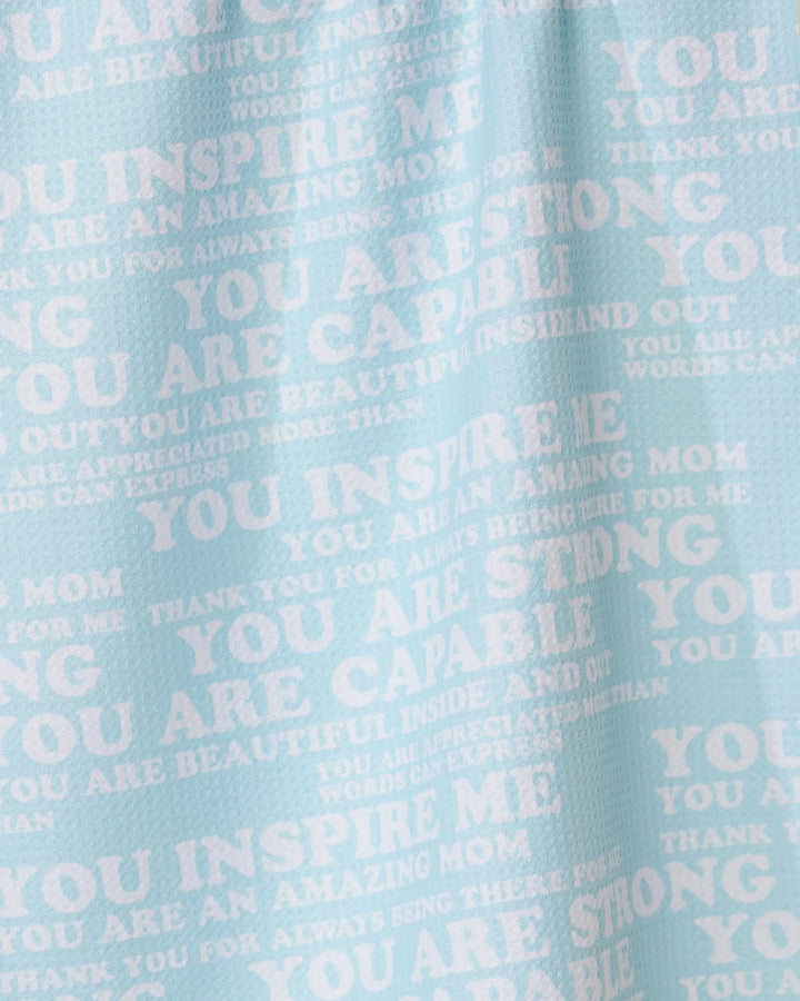 The GEOMETRY Mom Mantra tea towel is light blue, made from recycled materials, and features white overlapping motivational phrases like YOU INSPIRE ME and YOU ARE AMAZING MOM in different fonts and sizes.