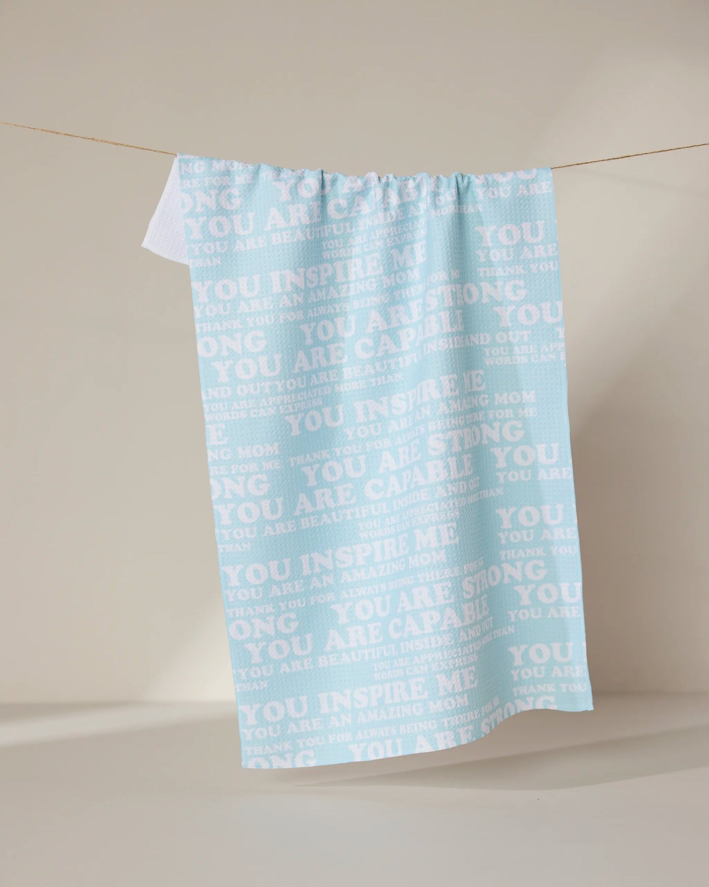 The GEOMETRY Mom Mantra is a light blue kitchen tea towel made from recycled materials, featuring white motivational phrases such as “You inspire me,” “You are strong,” and “You are capable.”.