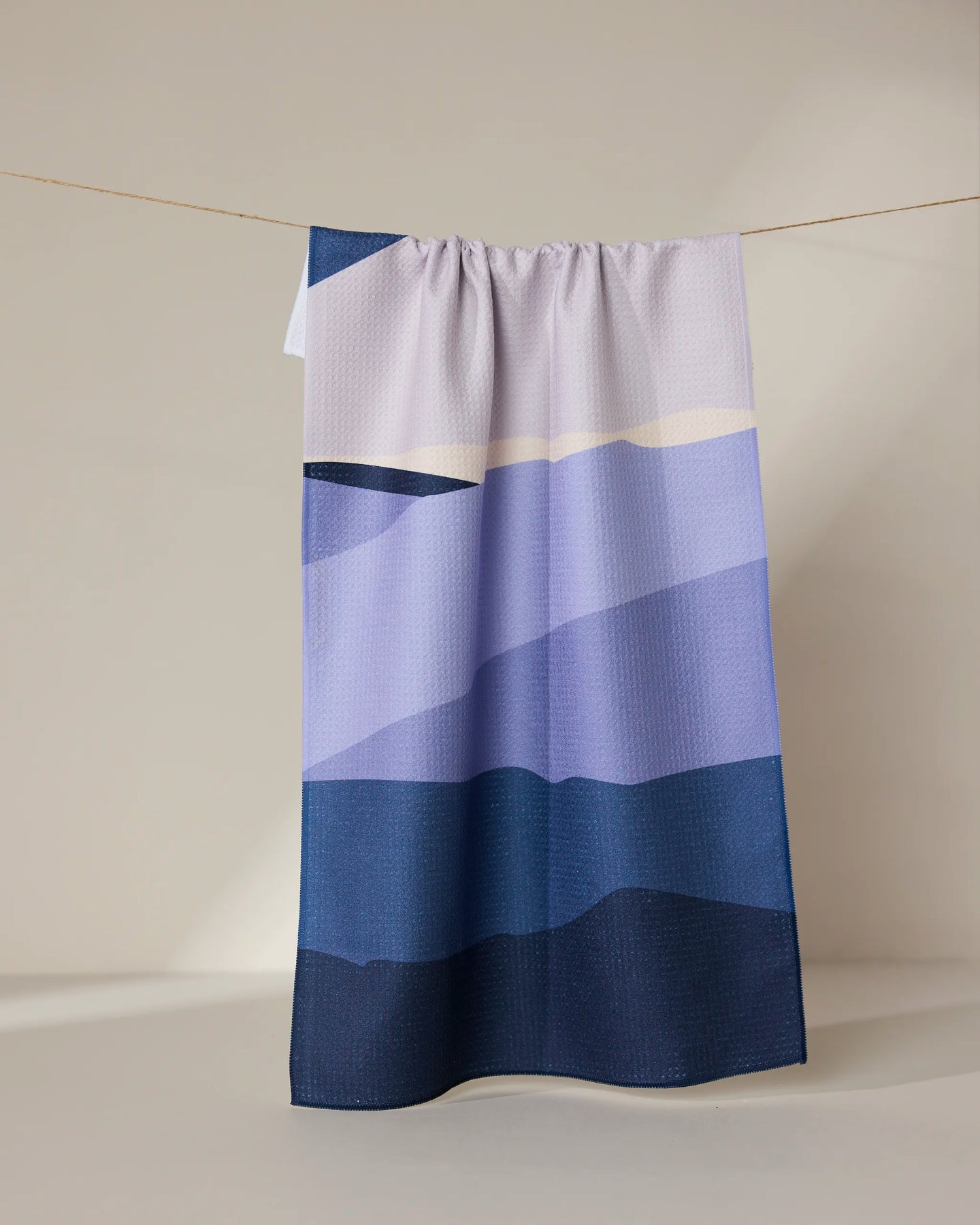 The GEOMETRY “Mountainous” tea towel, featuring absorbent fabric and abstract geometric shapes in blue, purple, and beige, hangs against a plain neutral background.