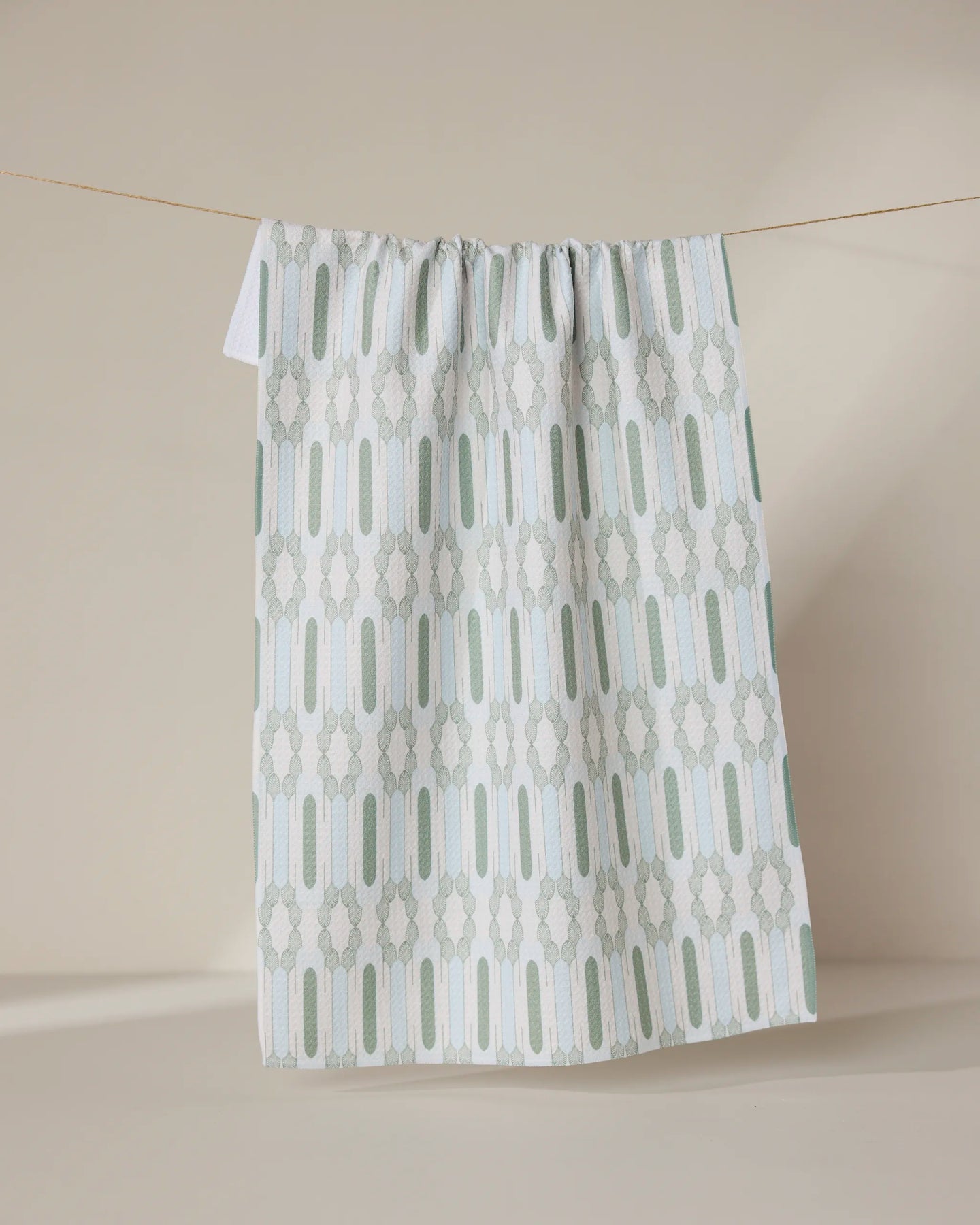 The GEOMETRY Musée Verte tea towel, made from recycled materials, showcases a light green and white geometric pattern with vertical and oval shapes, hanging on a string against a plain beige background.