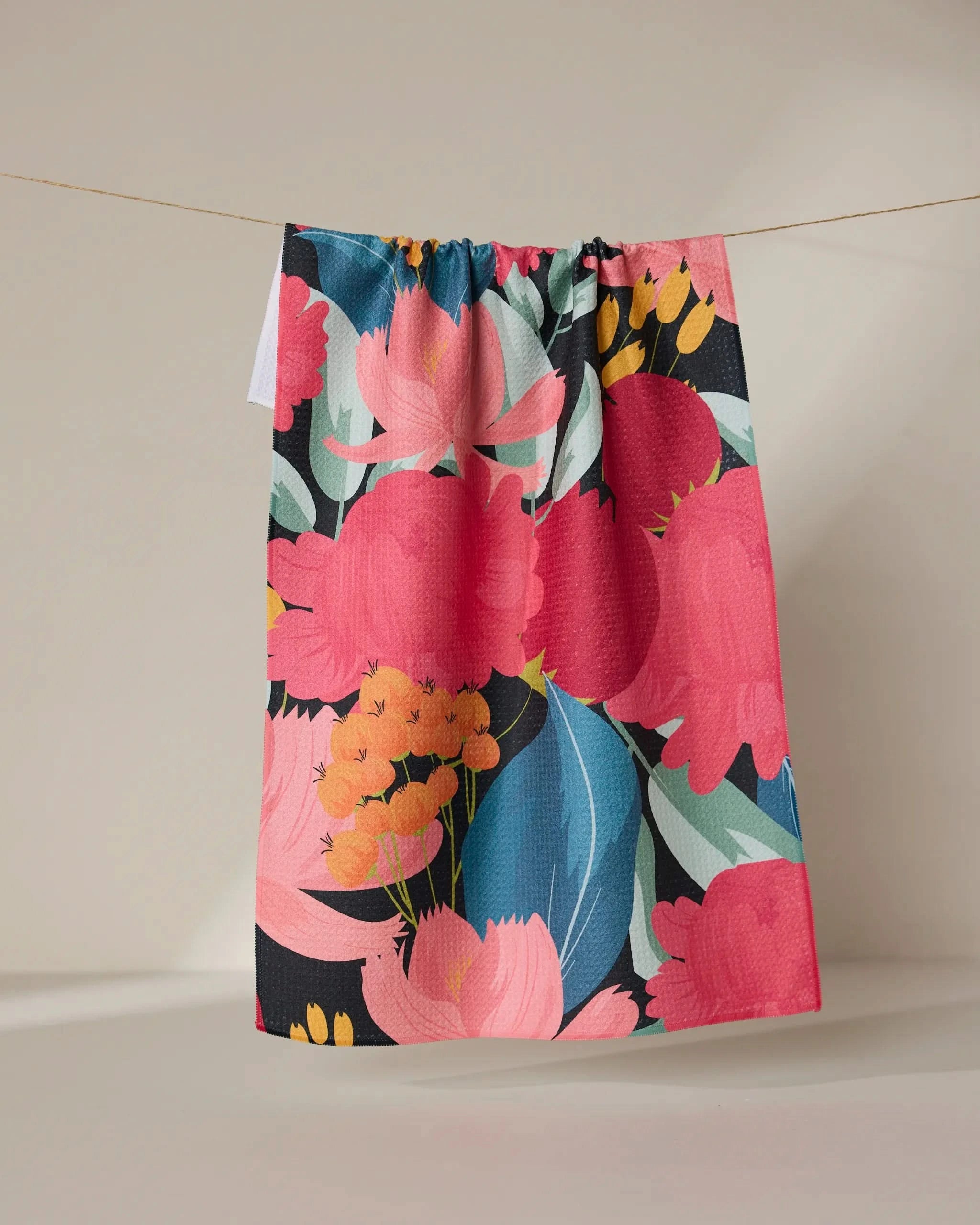 The GEOMETRY My Garden kitchen tea towel, featuring bold pink, red, blue, and yellow florals on a neutral beige background, hangs on a line—crafted with super-absorbent Geoweave technology.