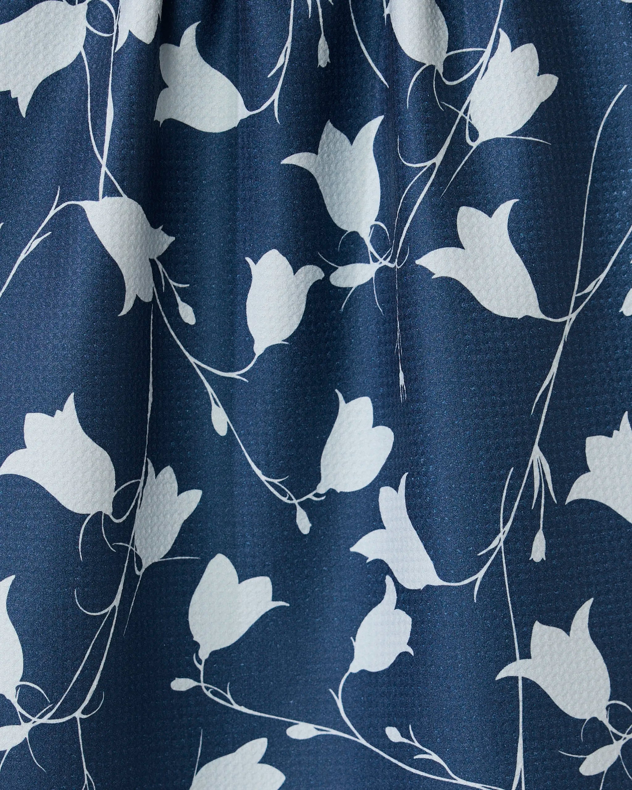 Close-up of GEOMETRYs Navy Dancing Flora recycled kitchen towel, showing a blue fabric with a white floral pattern of stylized flowers and thin, flowing stems on a textured surface with soft folds.