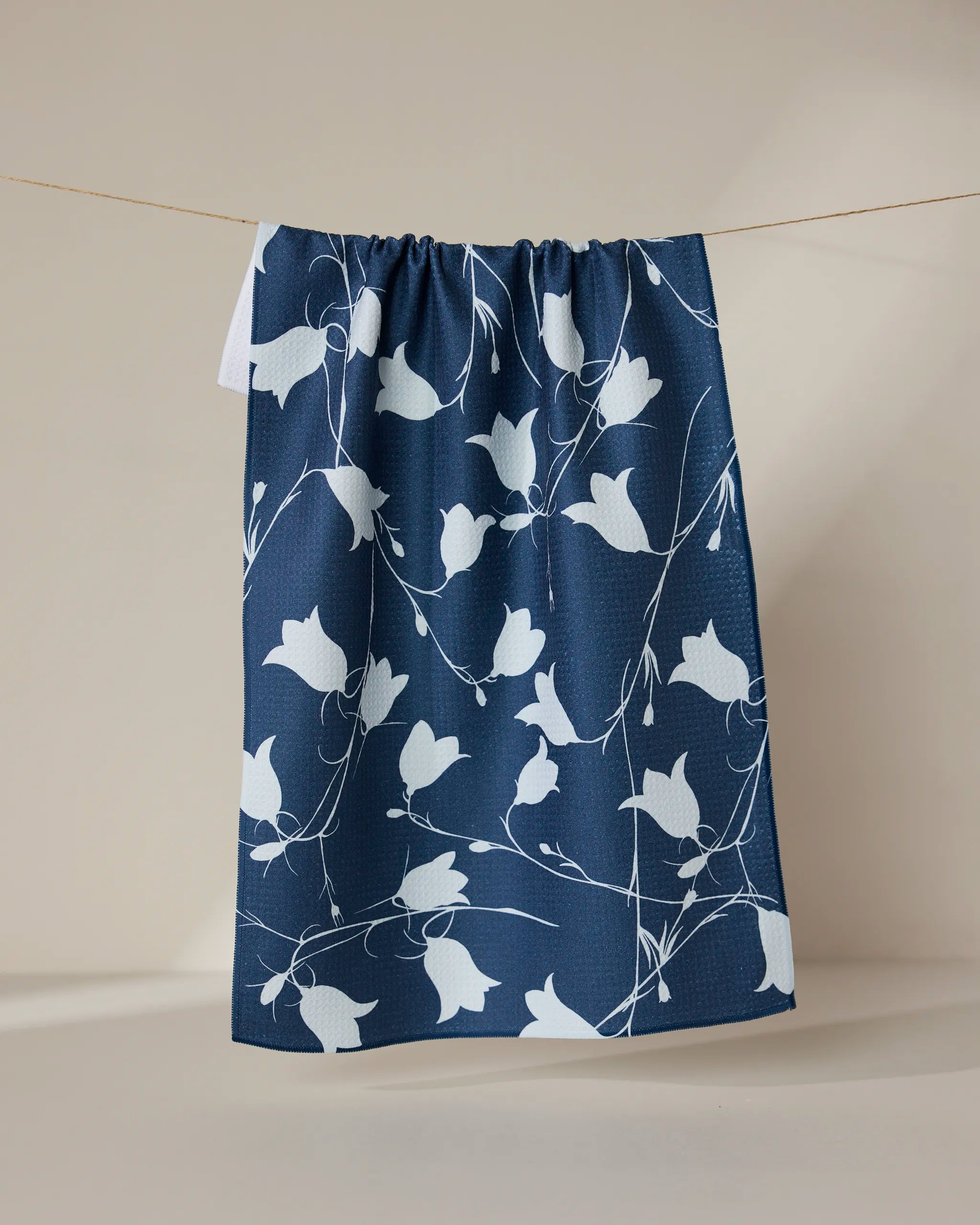 The GEOMETRY Navy Dancing Flora towel, featuring a white floral pattern on navy blue, hangs from a thin string against a plain beige background.