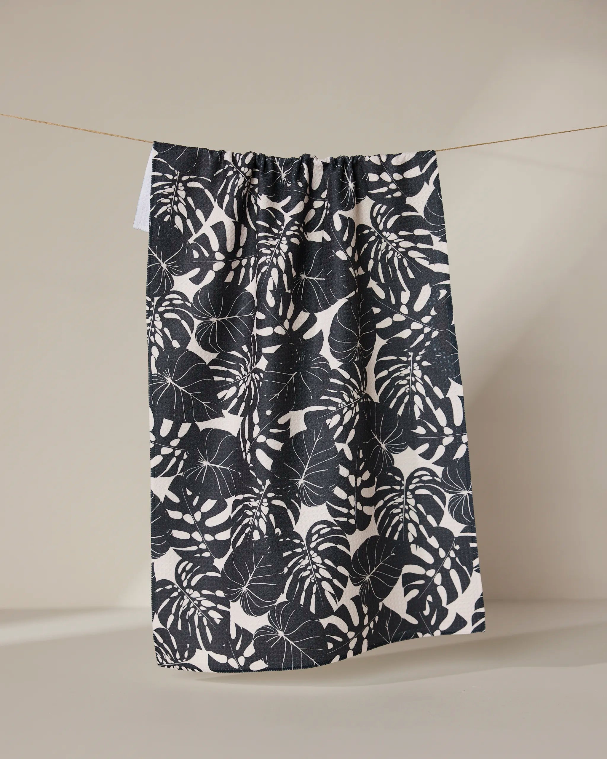 The GEOMETRY Neutral Leaves tea towel, featuring a bold black and white monstera leaf design and made from recycled materials, hangs on a clothesline against a plain beige background.