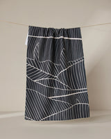 The GEOMETRY Neutral Palms tea towel, featuring a white geometric leaf pattern on black, hangs from a string against a plain beige background.