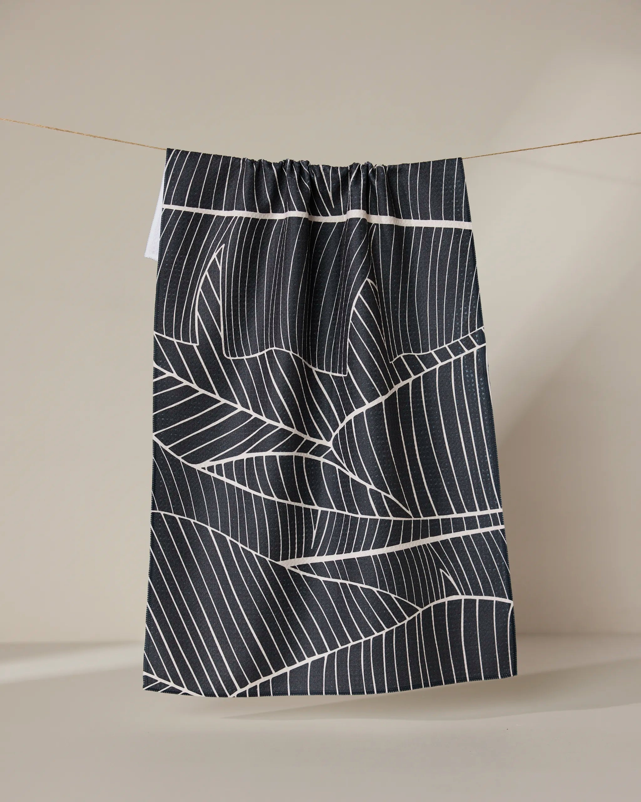The GEOMETRY Neutral Palms tea towel, featuring a white geometric leaf pattern on black, hangs from a string against a plain beige background.