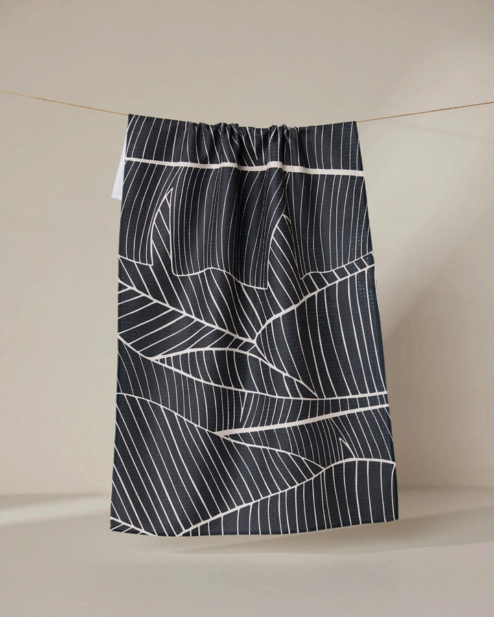 The GEOMETRY Neutral Palms tea towel, featuring a white geometric leaf pattern on black, hangs from a string against a plain beige background.