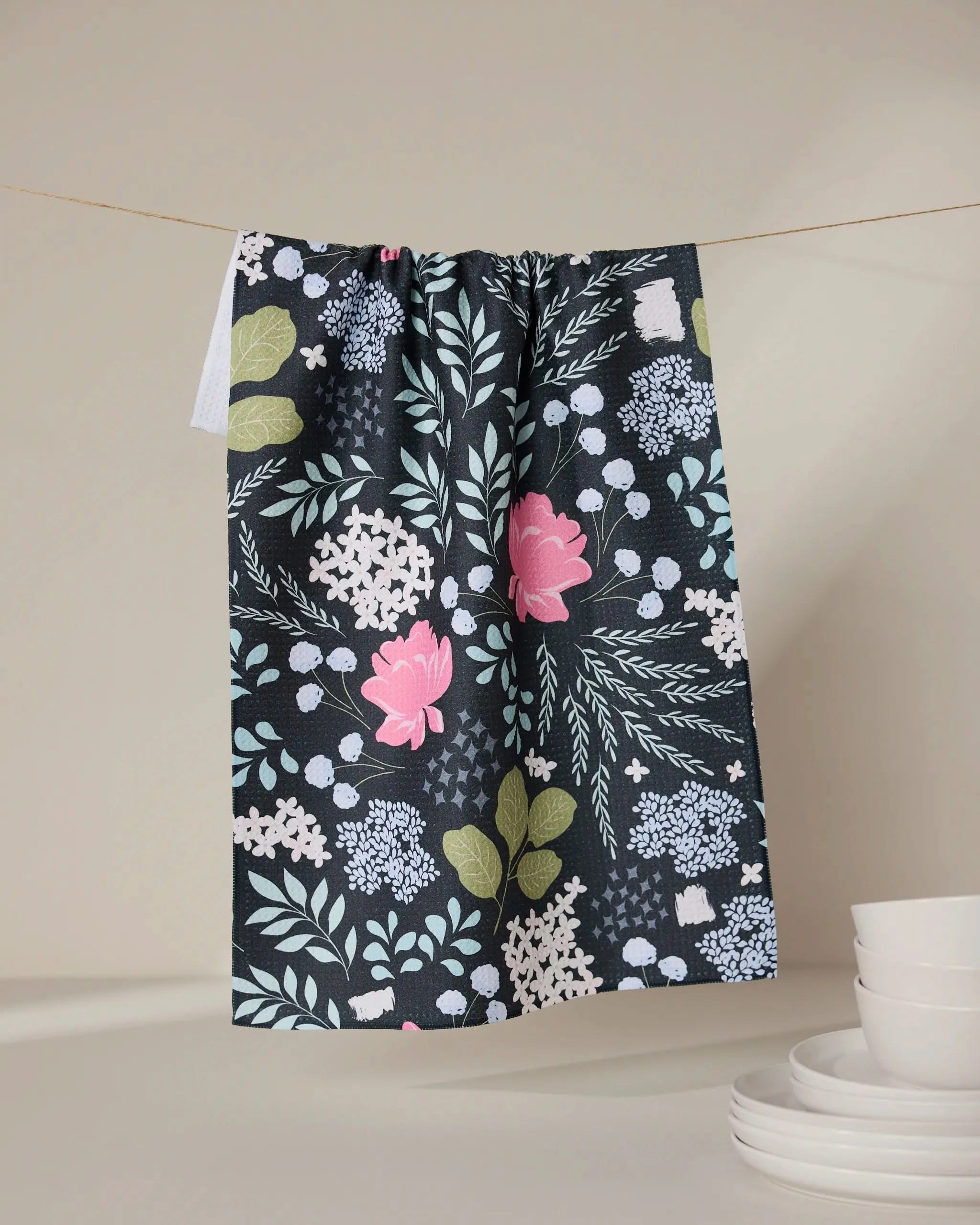 The GEOMETRY Not Forgotten recycled kitchen towel, featuring pink flowers, white blossoms, and green leaves on a dark background, hangs above a stack of white dishes against a neutral backdrop.