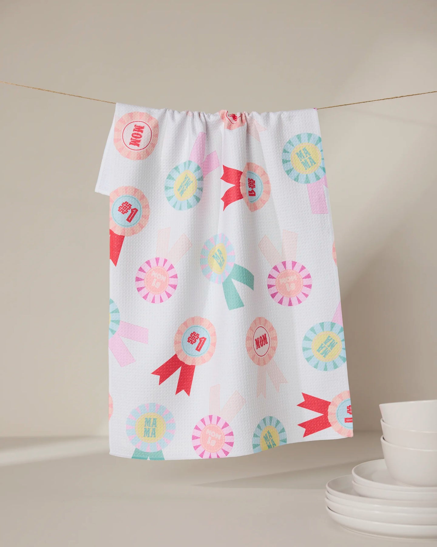 The “Number One Mom” tea towel by GEOMETRY, made from recycled materials, features colorful ribbon designs and “MOM” and “MAMA” text. It hangs above white dishes, offering an eco-friendly accent to a neutral kitchen.