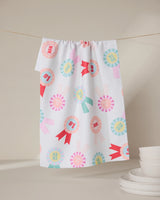 The “Number One Mom” tea towel by GEOMETRY, made from recycled materials, features colorful ribbon designs and “MOM” and “MAMA” text. It hangs above white dishes, offering an eco-friendly accent to a neutral kitchen.