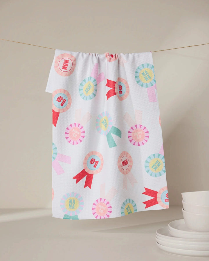 The “Number One Mom” tea towel by GEOMETRY, made from recycled materials, features colorful ribbon designs and “MOM” and “MAMA” text. It hangs above white dishes, offering an eco-friendly accent to a neutral kitchen.