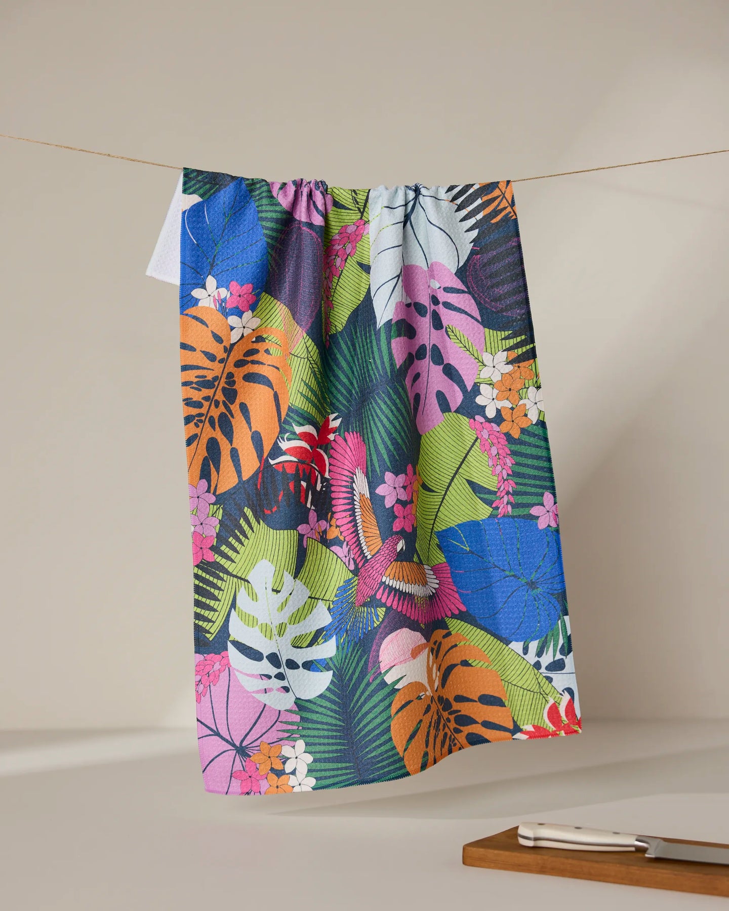 The GEOMETRY Palm Party recycled kitchen towel featuring a vibrant tropical leaf and flower pattern hangs on a clothesline, while a small wooden tray with a knife rests nearby against a neutral background.