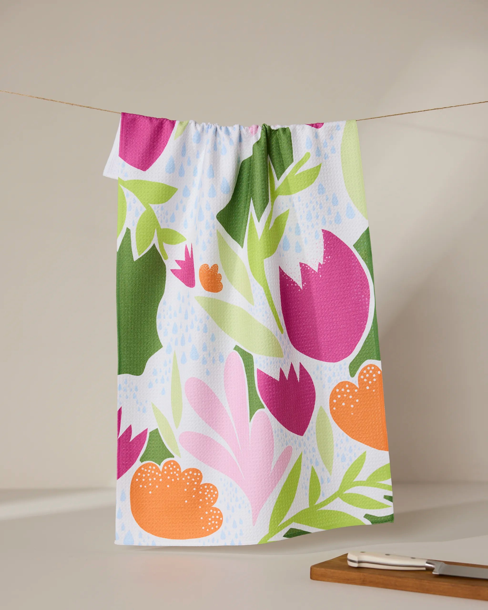 The GEOMETRY Paper Fields tea towel, featuring a vibrant floral pattern in pink, orange, green, and light blue, hangs on a string. Below on the countertop are a cutting board and knife.