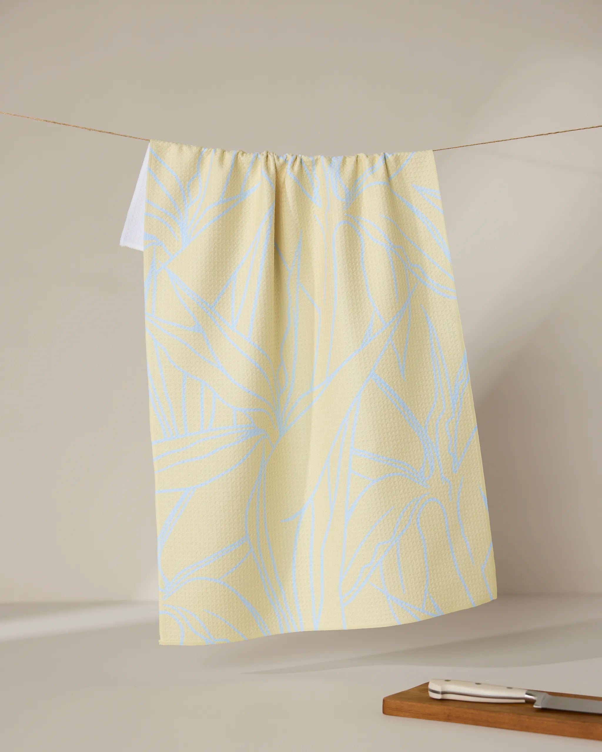 The GEOMETRY Paradise Blooms tea towel, featuring a light blue abstract leaf pattern on yellow, hangs on a line with a cutting board and knife placed nearby against a neutral background.
