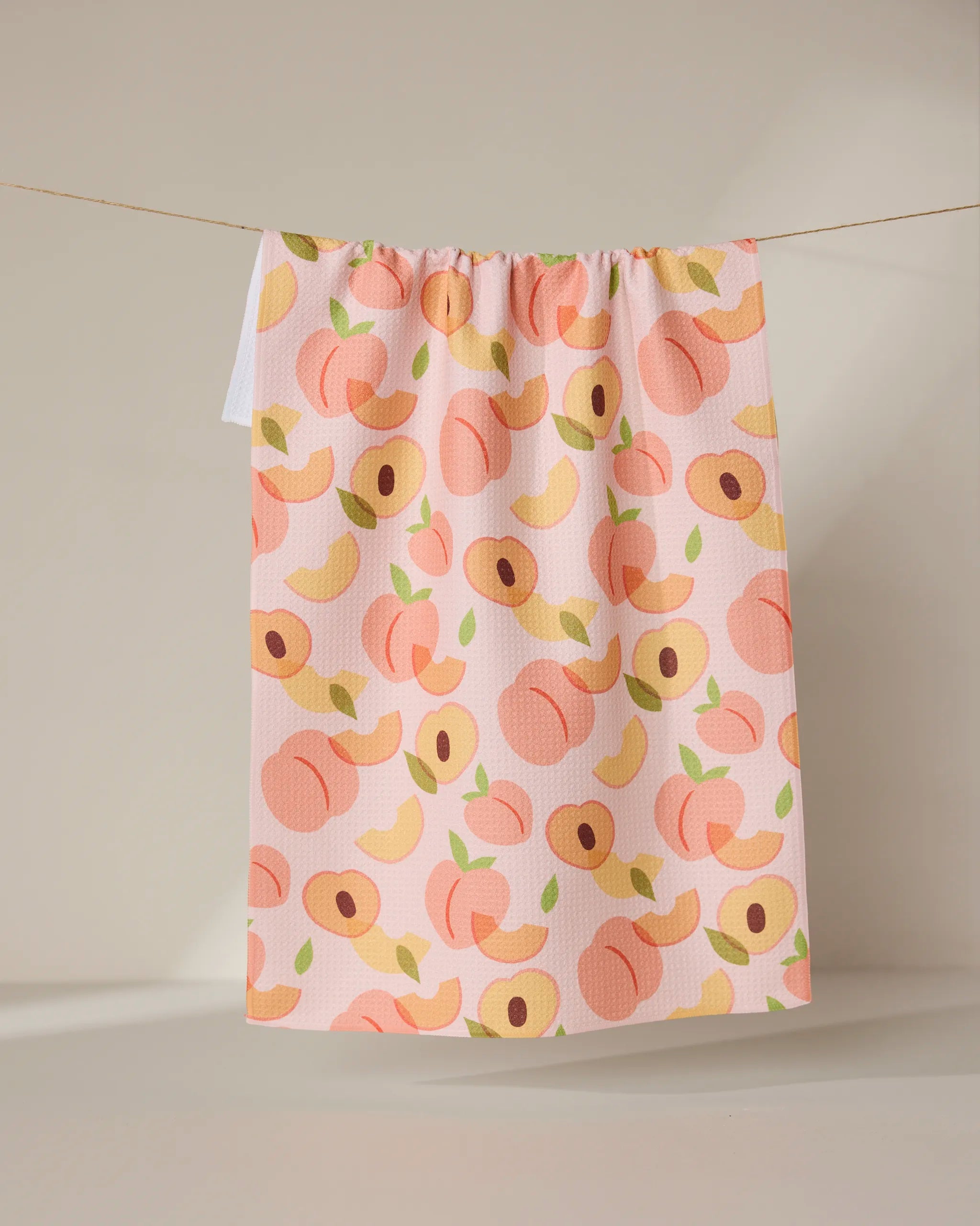 The GEOMETRY Peaches tea towel features playful whole and sliced peaches with green leaves in pink, orange, and yellow on a neutral background. Ultra-absorbent, it adds a cheerful pop of color to any kitchen.