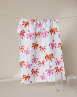 The GEOMETRY Pink Palms dish towel, featuring a vibrant pink and orange palm tree design, hangs on a line behind two clear ribbed glass cups set on a beige surface.