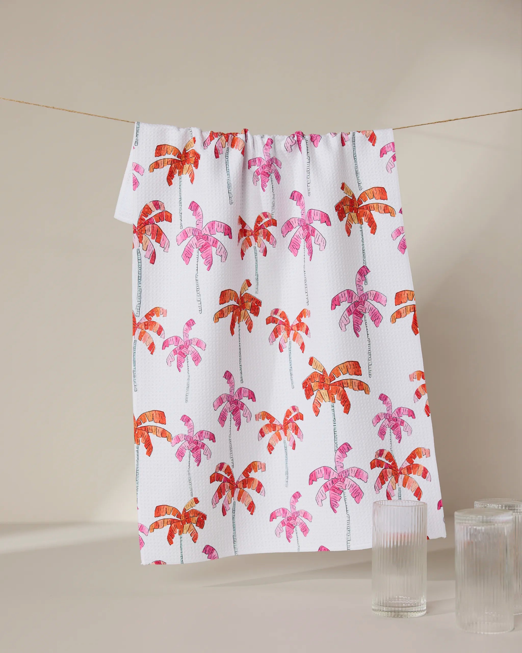 The GEOMETRY Pink Palms dish towel, featuring a vibrant pink and orange palm tree design, hangs on a line behind two clear ribbed glass cups set on a beige surface.