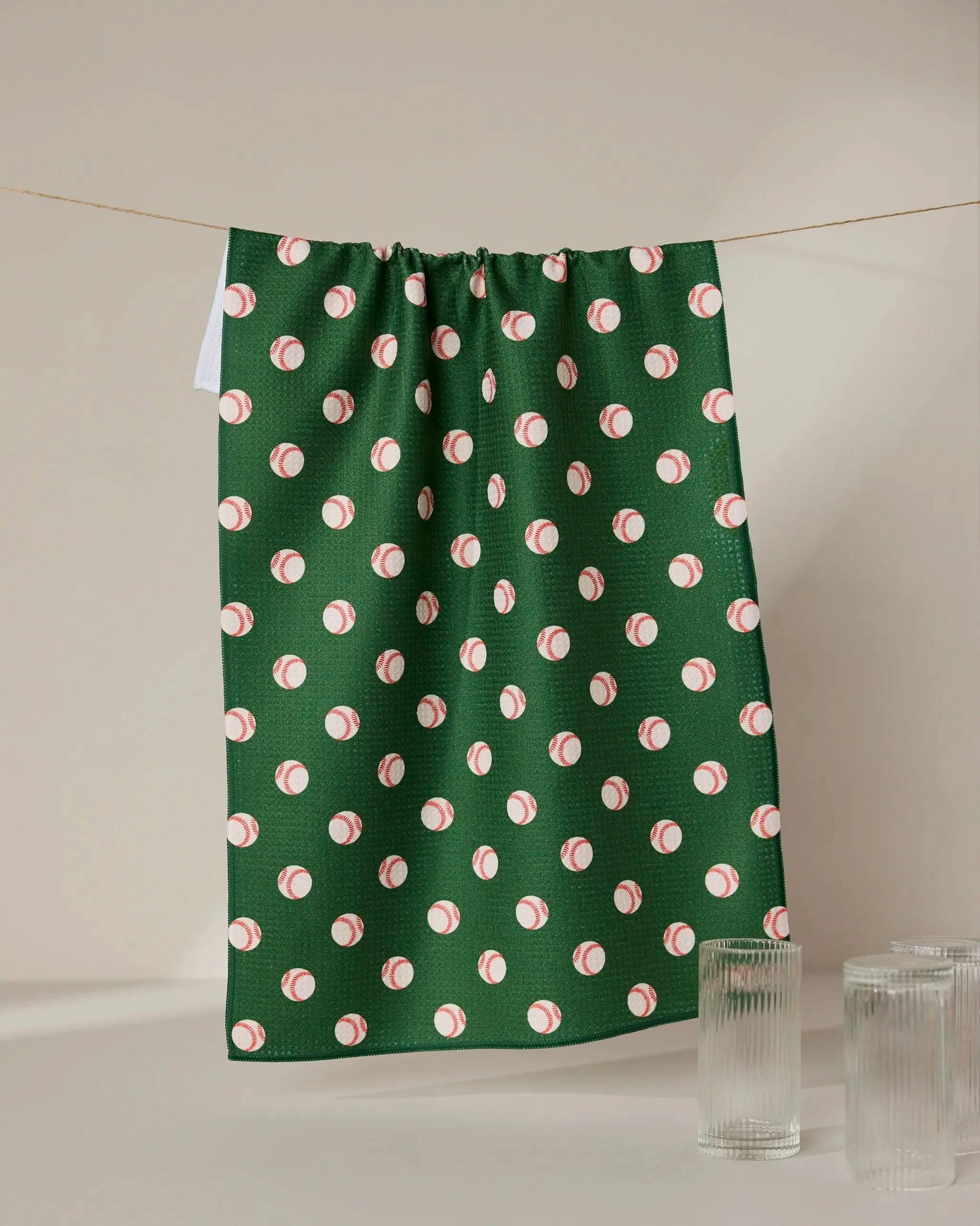 The GEOMETRY Play Ball kitchen towel, featuring a green hue and a repeating small baseball pattern, hangs on a string between two glass cups against a simple light background.
