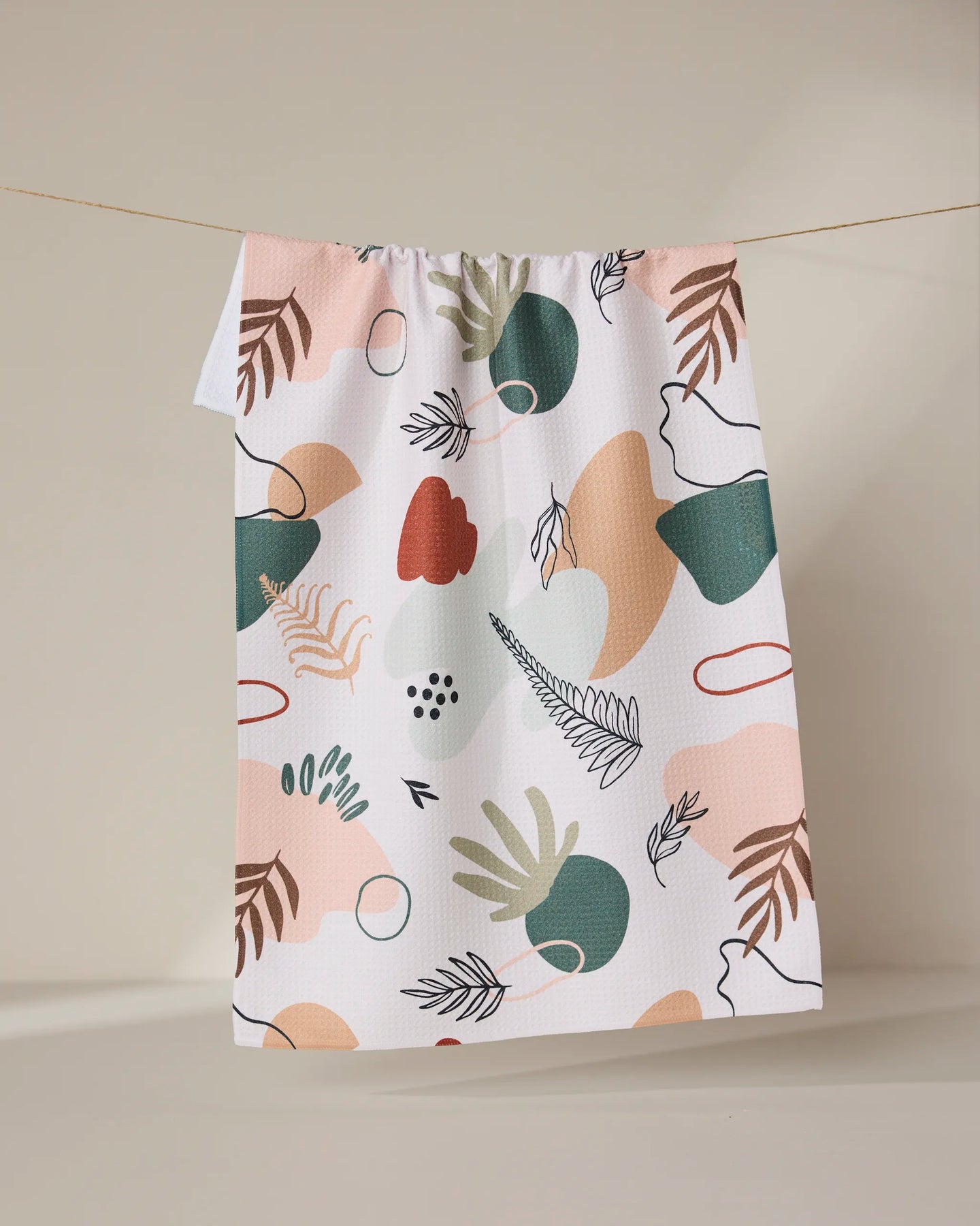 The GEOMETRY Plated Ferns tea towel, made from recycled materials, showcases an abstract leaf pattern in green, pink, beige, and red hues. Quick-drying and vibrant, it’s pictured hanging on a clothesline against a neutral backdrop.