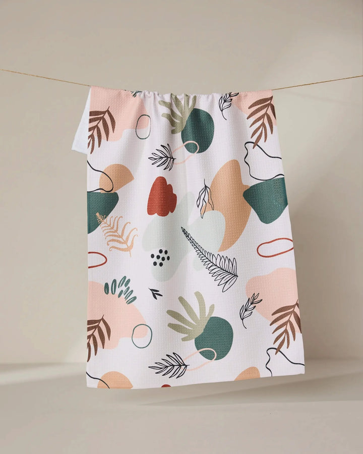 The GEOMETRY Plated Ferns tea towel, made from recycled materials, showcases an abstract leaf pattern in green, pink, beige, and red hues. Quick-drying and vibrant, it’s pictured hanging on a clothesline against a neutral backdrop.