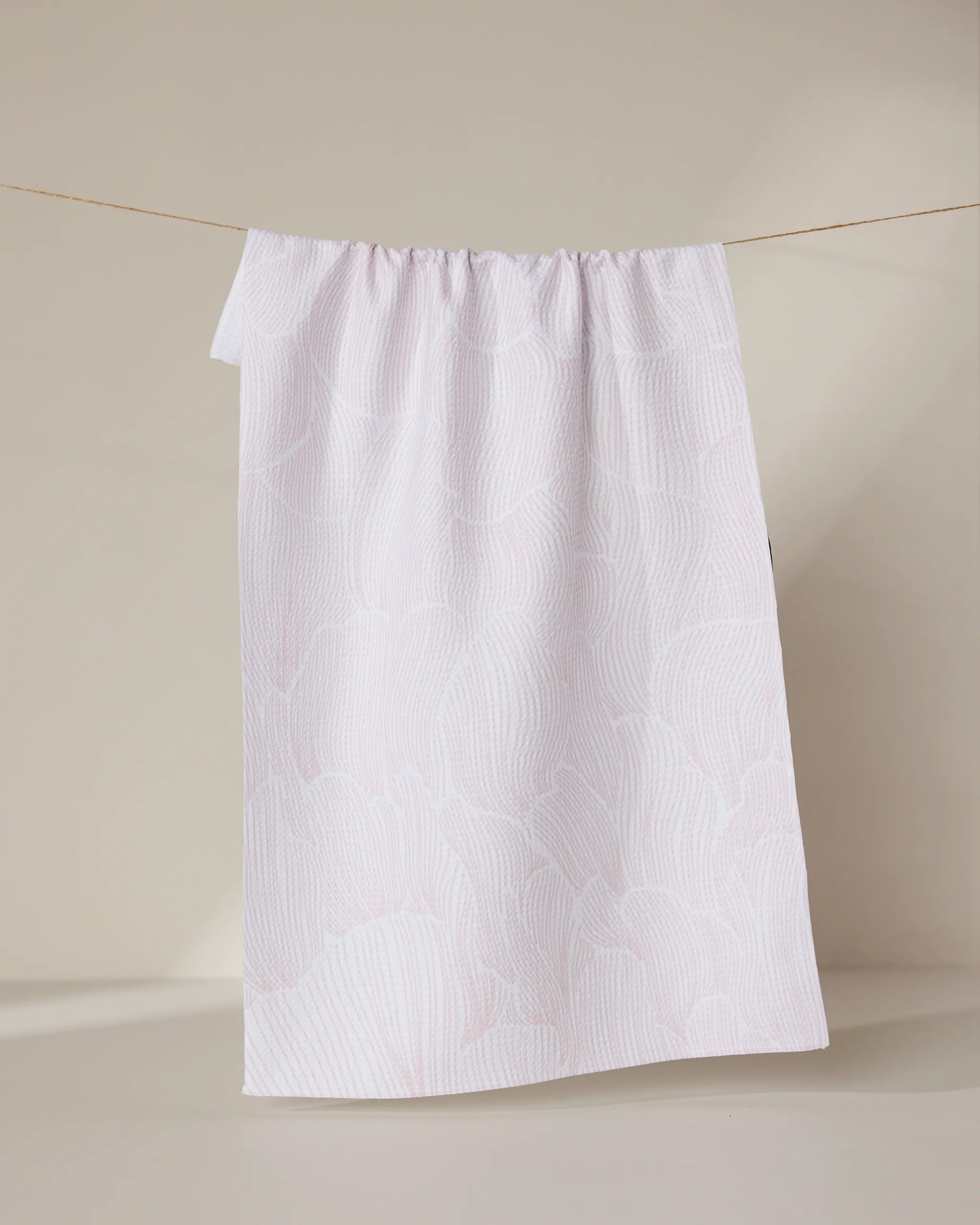 The Posh Petals Blushed Bergamot tea towel by Geometry, featuring a pale pink wavy pattern and crafted from recycled materials, hangs over a thin string against a neutral beige background.
