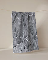 The Geometry Posh Petals Fresh Linen tea towel, featuring black and white abstract wavy lines, hangs on a string against a plain beige background.