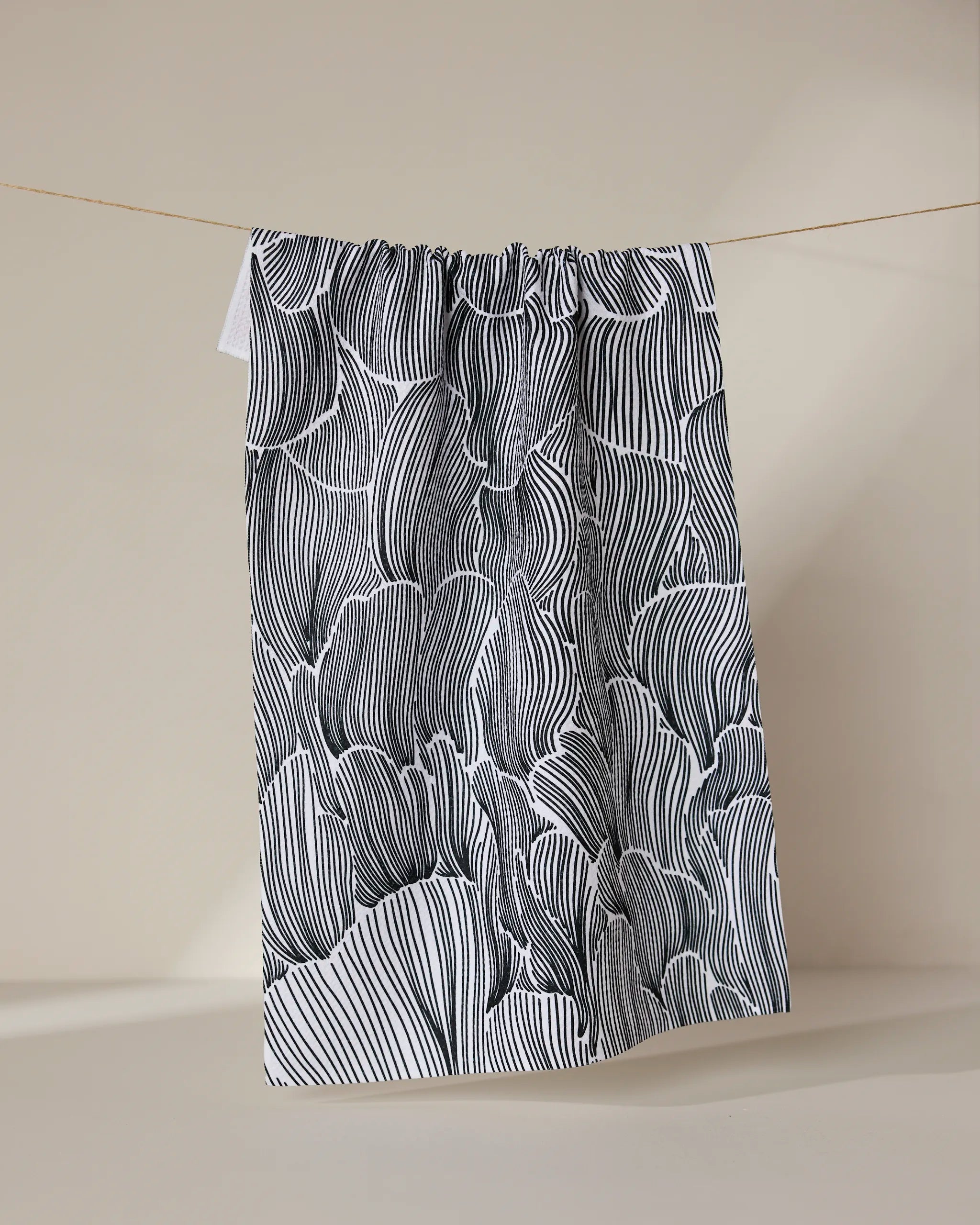 The Geometry Posh Petals Fresh Linen tea towel, featuring black and white abstract wavy lines, hangs on a string against a plain beige background.