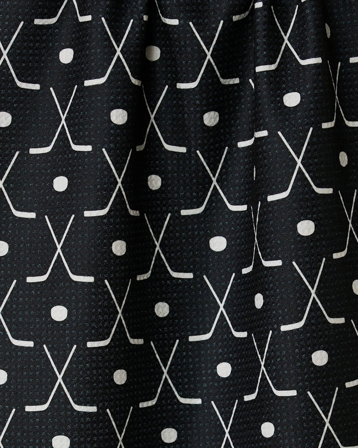 Close-up of the GEOMETRY Puck tea towel with ultra-absorbent black fabric, featuring a repeating grid pattern of white crossed hockey sticks and pucks for a sporty look.