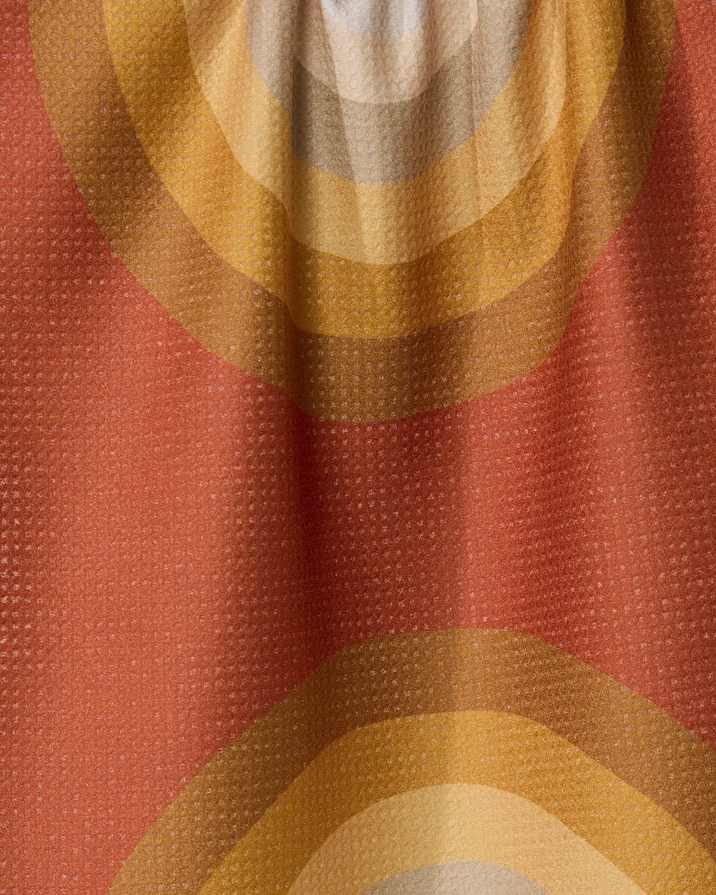 A close-up of GEOMETRYs Pumpkin Spice fabric shows textured, recycled material with concentric circles in orange, yellow, and beige on a muted red background, highlighted by visible folds and shadows.
