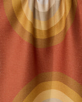 A close-up of GEOMETRYs Pumpkin Spice fabric shows textured, recycled material with concentric circles in orange, yellow, and beige on a muted red background, highlighted by visible folds and shadows.