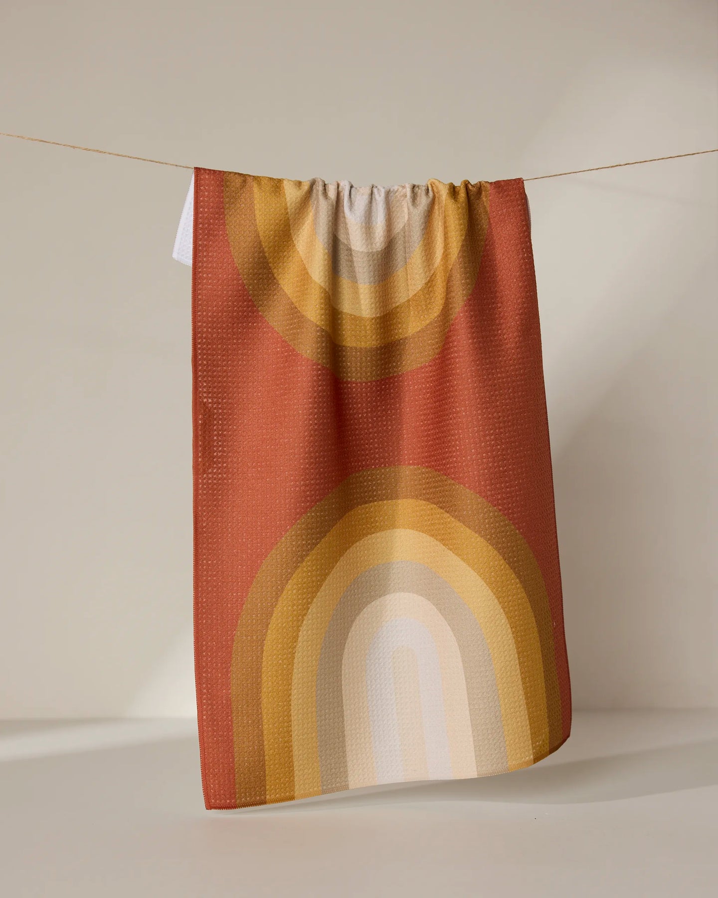 The GEOMETRY “Pumpkin Spice” towel, featuring a retro rainbow pattern in warm orange, yellow, and beige tones, hangs on a thin string against a neutral background.