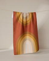 The GEOMETRY “Pumpkin Spice” towel, featuring a retro rainbow pattern in warm orange, yellow, and beige tones, hangs on a thin string against a neutral background.