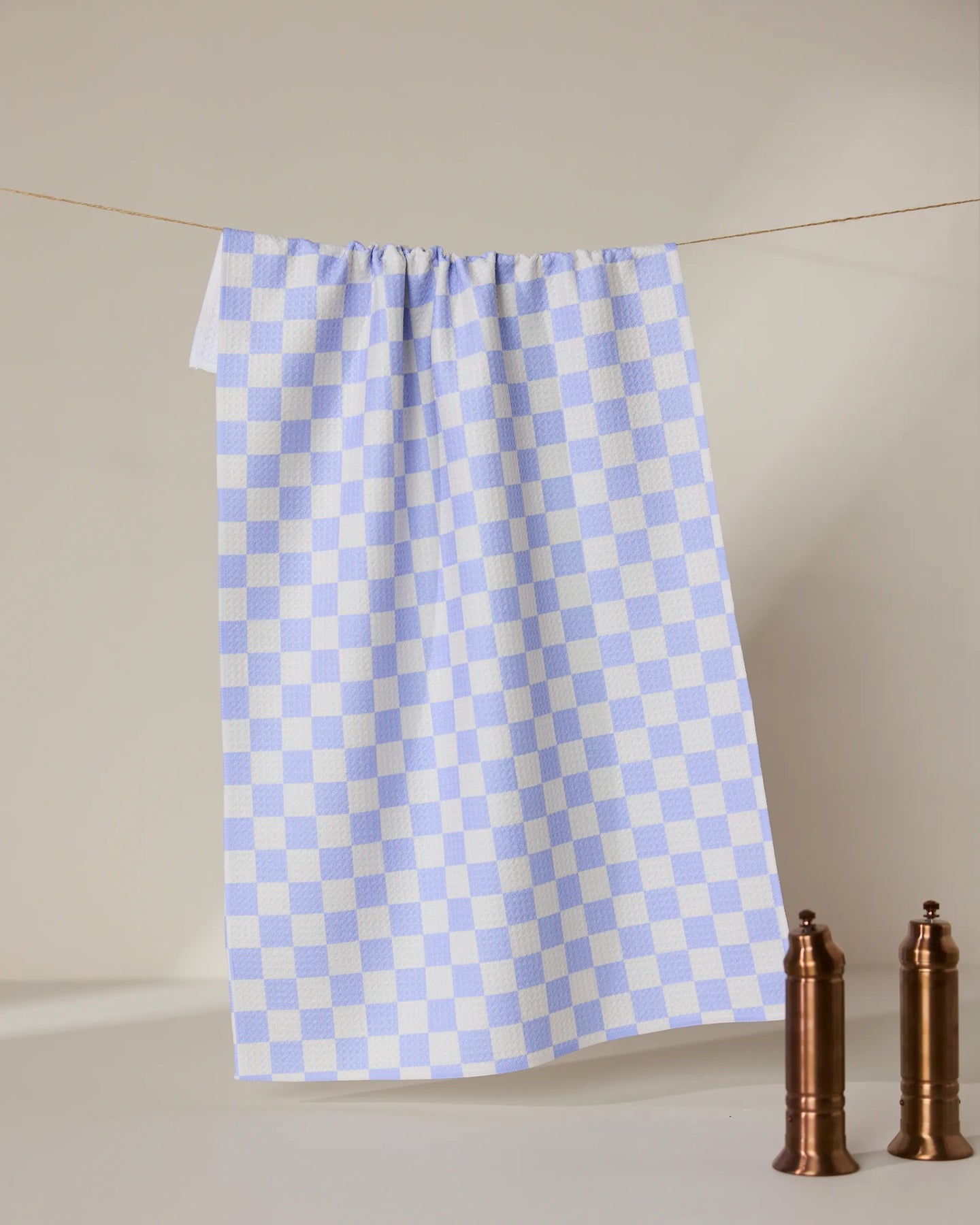 A Purple Picnic recycled kitchen towel by GEOMETRY hangs on a string against a beige background, with two brown mills on a white surface in the lower right corner.