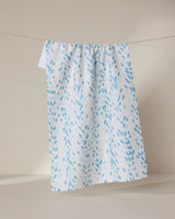 The GEOMETRY Reeds Printed - Midday is a white tea towel featuring light blue leaf-like patterns, crafted from recycled materials, shown hanging neatly on a clothesline against a neutral background.